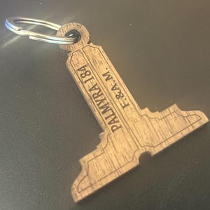 Puede incluir: Un llavero de madera con un llavero plateado. El llavero tiene forma de llave estilizada y presenta el texto grabado "PALMYRA 184 F.&A.M.". La madera tiene una veta natural y un color marrón cálido.