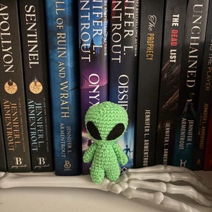 May include: A small, green crocheted alien figure with black eyes stands on a white, skeletal hand. The background features a shelf lined with books by Jennifer L. Armentrout, with titles like "Fall of Ruin and Wrath" and "Onyx."