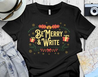 May include: Black t-shirt with a Christmas-themed design. The design features a wreath with holly, bells, and presents. The text "Be Merry & Write" is in the center of the wreath.