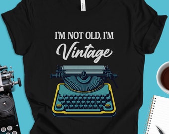 May include: Black t-shirt with the text "I'M NOT OLD, I'M Vintage" above a teal and yellow vintage typewriter illustration. The shirt is made of a soft material and is suitable for casual wear.