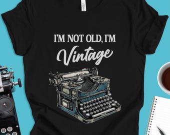 May include: Black t-shirt with the text "I'M NOT OLD, I'M Vintage" in white script above a detailed illustration of a vintage typewriter in shades of blue and gray.
