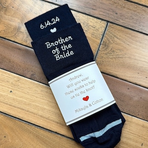 May include: A pair of dark blue socks with white embroidery that reads "Brother of the Bride" and "6.14.24". A white card with red heart and text that reads "Andrew, Will you wear these socks to help us tie the knot? Mikayla & Colton" is attached to the socks.