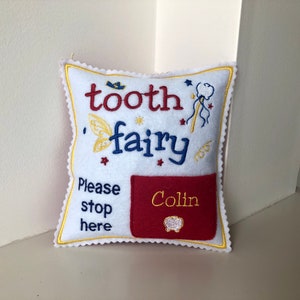 May include: A white square felt pillow with scalloped edges, embroidered with the text "tooth fairy" in red and blue, "Please stop here" in blue, and a red pocket featuring the name "Colin" in yellow thread and a small tooth icon.