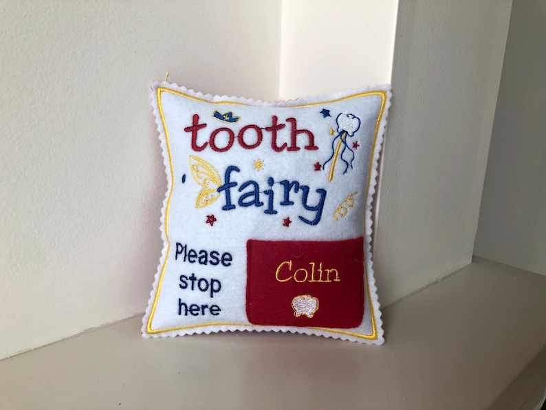 May include: A white square felt pillow with scalloped edges, embroidered with the text "tooth fairy" in red and blue, "Please stop here" in blue, and a red pocket featuring the name "Colin" in yellow thread and a small tooth icon.