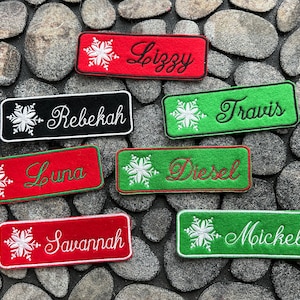 May include: Rectangular embroidered patches in red, black, and green, each with a name in cursive script and a snowflake design. Names include Lizzy, Rebekah, Travis, Luna, Diesel, Savannah, and Mickel. Patches are on a stone background.