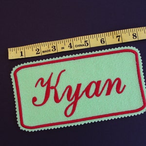 May include: A light green felt name patch with a red border and the name "Kyan" embroidered in red script. The patch is approximately 5 inches long. A yellow measuring tape is placed above the patch.
