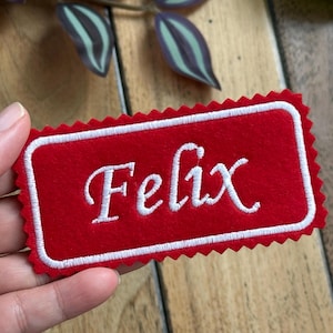 May include: Red felt name tag with a white border and the name "Felix" embroidered in white.