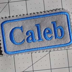 May include: A gray felt name tag with a blue border and the name "Caleb" embroidered in blue.