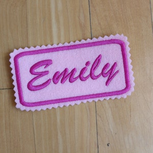 May include: A pink felt name patch embroidered with the name "Emily" in a darker pink script. The patch is rectangular with a scalloped edge.