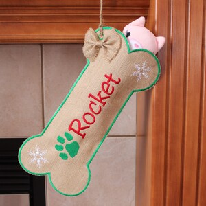 May include: A beige burlap dog bone-shaped Christmas stocking with the name "Rocket" in red script. It features a green paw print, snowflakes, and a green border. A small burlap bow and a pink pig peek out from behind the stocking.