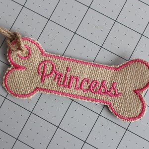 May include: A pink embroidered dog bone tag with the word "Princess" on it. The tag is made of burlap and has a twine loop for attaching to a collar.