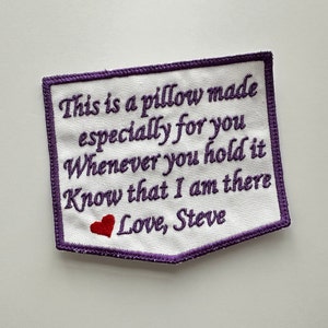 May include: Purple embroidered patch with a white fabric background. The text reads "This is a pillow made especially for you. Whenever you hold it know that I am there. Love, Steve". A small red heart is embroidered below the text.