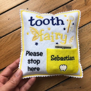 May include: A white felt square pillow with yellow scalloped edges, embroidered with the text "tooth fairy" in blue and yellow, "Please stop here" in black, and a yellow pocket featuring the name "Sebastian" and a white tooth icon.
