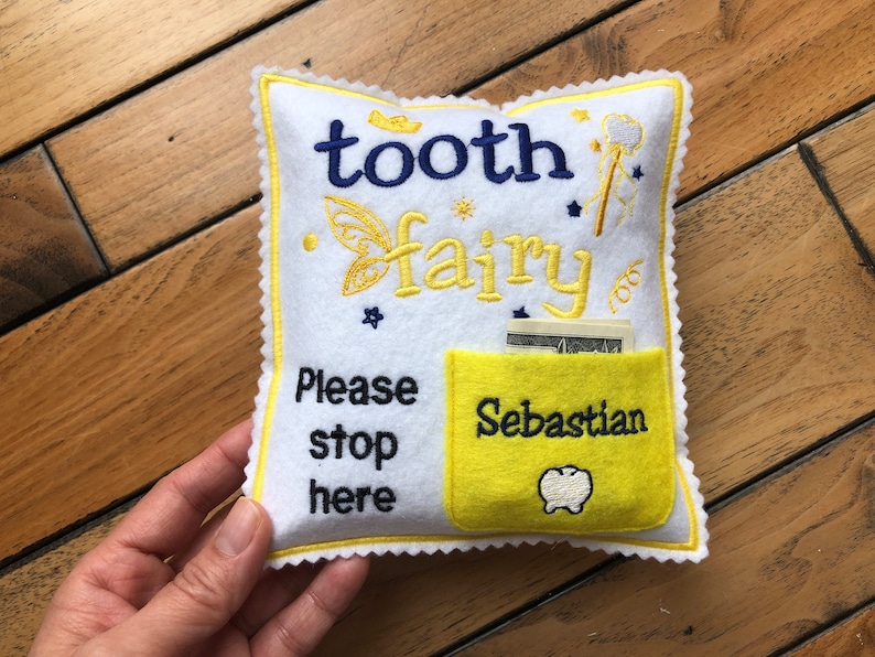 May include: A white felt square pillow with yellow scalloped edges, embroidered with the text "tooth fairy" in blue and yellow, "Please stop here" in black, and a yellow pocket featuring the name "Sebastian" and a white tooth icon.