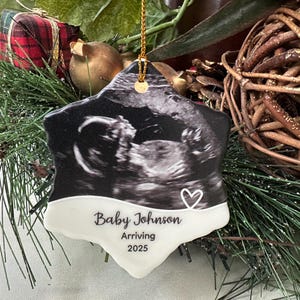 May include: A star-shaped Christmas ornament featuring a black and white sonogram image. The ornament has a white bottom section with the text "Baby Johnson Arriving 2025" and a heart symbol. A gold string is attached for hanging.