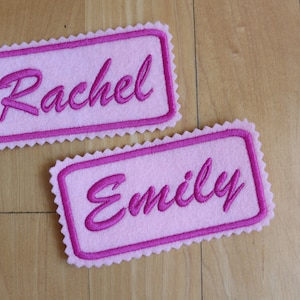 May include: Two pink felt name tags with pink stitching. The top tag says "Rachel" and the bottom tag says "Emily".