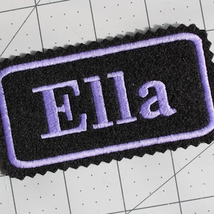 May include: Purple embroidered name tag with the name "Ella" on a black background.