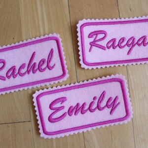 May include: Three pink felt name tags with pink stitching. The names are "Rachel", "Raegan", and "Emily".
