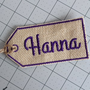 May include: A brown burlap gift tag with a purple stitched border and the name "Hanna" embroidered in purple thread.