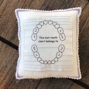 May include: A square white felt pillow with a purple stitched border. The center features a printed graphic of a tooth chart on lined paper with the text, This lost-tooth chart belongs to, and a blank line for writing a name.