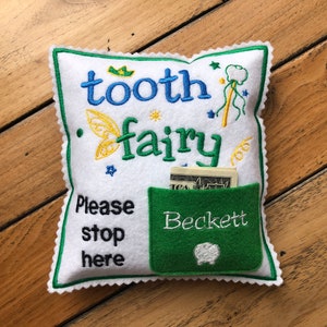 May include: A white square felt pillow with scalloped green edges, embroidered with the text "tooth fairy" and "Please stop here". It features a green pocket with the name "Beckett" and a small tooth icon, containing a US dollar bill.