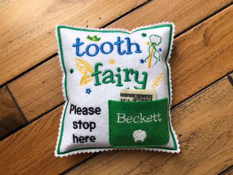 May include: A white square felt pillow with scalloped green edges, embroidered with the text "tooth fairy" and "Please stop here". It features a green pocket with the name "Beckett" and a small tooth icon, containing a US dollar bill.