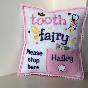 May include: A white square felt pillow with pink scalloped edges, embroidered with the text "tooth fairy" and "Please stop here". It features a yellow fairy wing, a crown, a wand, and a pink pocket labeled "Hailey" with a tooth icon.