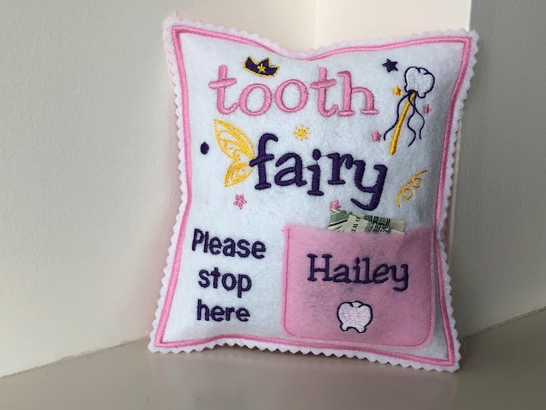 May include: A white square felt pillow with pink scalloped edges, embroidered with the text "tooth fairy" and "Please stop here". It features a yellow fairy wing, a crown, a wand, and a pink pocket labeled "Hailey" with a tooth icon.