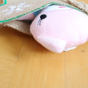 May include: A pink plush toy seal peeking out of a burlap sack with a green trim. The seal has black eyes and whiskers. The sack has a decorative embroidered design.