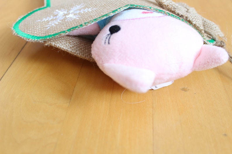 May include: A pink plush toy seal peeking out of a burlap sack with a green trim. The seal has black eyes and whiskers. The sack has a decorative embroidered design.