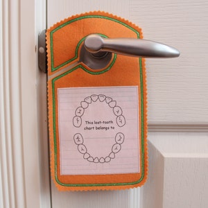 May include: Orange felt door hanger with a green border and a white paper insert. The insert has a drawing of a circle of teeth and the text "This lost-tooth chart belongs to".