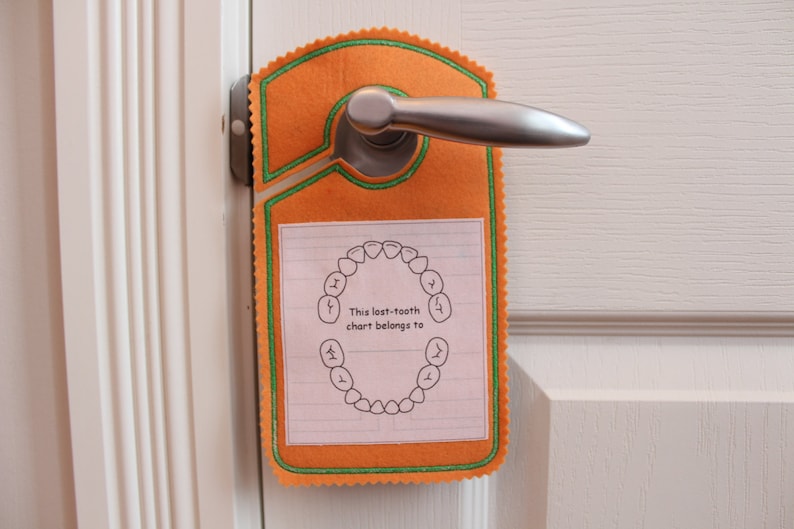 May include: Orange felt door hanger with a green border and a white paper insert. The insert has a drawing of a circle of teeth and the text "This lost-tooth chart belongs to".