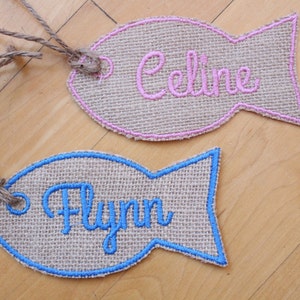 May include: Two burlap fish-shaped gift tags with blue and pink embroidered edges. The tag on the left has the name "Flynn" embroidered in blue thread. The tag on the right has the name "Celine" embroidered in pink thread.