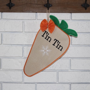 May include: A carrot-shaped fabric decoration with an orange bow and green leaves. The beige carrot has an orange outline and the text "Tin Tin" with a snowflake. It hangs from a wooden hook against a white brick wall.