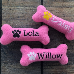 May include: Three pink plush dog bone toys with embroidered paw prints and names: Lola, Daisy, and Willow.