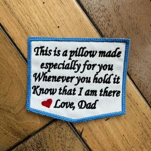 May include: White fabric pillow with blue edging and embroidered text: "This is a pillow made especially for you. Whenever you hold it know that I am there. Love, Dad." A red heart is embroidered next to the text "Love, Dad."