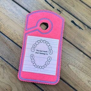 May include: Pink felt tooth chart with a hole at the top for hanging. The chart has a circle with teeth drawn inside and the text "This lost-tooth chart belongs to".