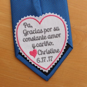 May include: Blue tie with a white heart-shaped patch. The patch has a pink scalloped border and features embroidered text in gray and a pink heart. The text reads "Pa, Gracias por su constante amor y cariño. Christine 6.17.17."