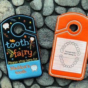 May include: Two tooth fairy door hangers. One is blue with black and yellow embroidery that says "tooth fairy" and "Please stop here for Maddox's tooth". The other is orange with white embroidery that says "This lost-tooth chart belongs to".