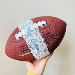 May include: A brown and white football with white lace and blue text that says "Nice Catch!" The football is labeled "National Football Conference" and "NFL League".