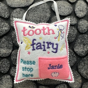 May include: A white felt square pillow with a pink scalloped edge and a white hanging ribbon. The pillow features embroidered text reading tooth fairy and Please stop here, along with a pink pocket embroidered with the name Janie.