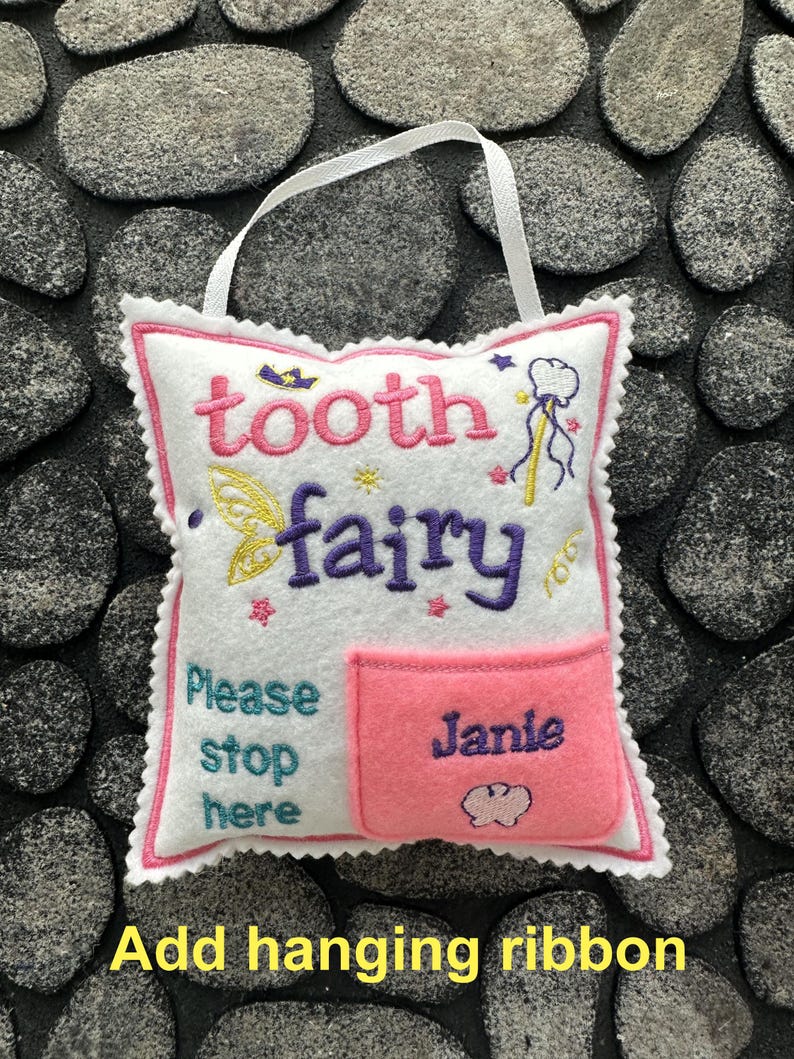 May include: A white felt square pillow with a pink scalloped edge and a white hanging ribbon. The pillow features embroidered text reading tooth fairy and Please stop here, along with a pink pocket embroidered with the name Janie.