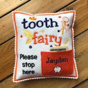 May include: A white square felt pillow with orange trim featuring the embroidered text tooth fairy, Please stop here, and the name Jayden. It includes a small orange pocket containing paper currency and embroidered icons of a crown, wand, and tooth.
