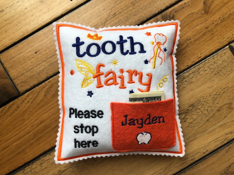 May include: A white square felt pillow with orange trim featuring the embroidered text tooth fairy, Please stop here, and the name Jayden. It includes a small orange pocket containing paper currency and embroidered icons of a crown, wand, and tooth.