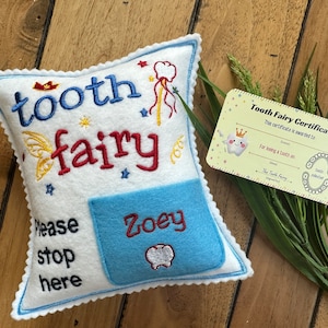 May include: A white felt pillow with blue scalloped edges, embroidered with the text "tooth fairy" and "Please stop here". A light blue pocket features the name "Zoey" and a tooth icon. Includes a "Tooth Fairy Certificate" card.