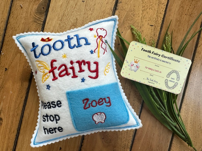 May include: A white felt pillow with blue scalloped edges, embroidered with the text "tooth fairy" and "Please stop here". A light blue pocket features the name "Zoey" and a tooth icon. Includes a "Tooth Fairy Certificate" card.