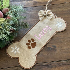 May include: A burlap dog bone-shaped Christmas ornament with the name "Lucy" in pink script. It features a brown paw print, white snowflakes, and a small burlap bow. The ornament hangs from a natural twine.