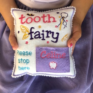 May include: A white felt square pillow with scalloped edges, embroidered with the text tooth fairy, a wand, and wings. It features a purple pocket with the name Celine and a tooth icon, plus the blue embroidered text Please stop here.