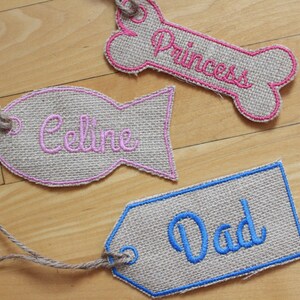 May include: Three burlap dog tags with pink, blue, and pink embroidery. The tags are shaped like a bone, a fish, and a rectangle. The text on the tags reads "Princess", "Celine", and "Dad".