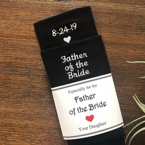 May include: Black socks with white embroidery that reads "Father of the Bride" and "8-24-19". A white heart is embroidered above the date. A white tag attached to the socks reads "Especially for the Father of the Bride" with a red heart and the text "Your Daughter".